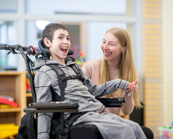 Eine Betreuerin hält die Hand eines lachenden Jungen der im Rollstuhl sitzt | © FatCamera - Getty Images