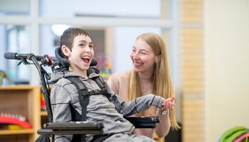 Eine Betreuerin hält die Hand eines lachenden Jungen der im Rollstuhl sitzt | © FatCamera - Getty Images